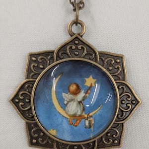 Peut inclure: Un collier de couleur bronze avec un pendentif sur le th&egrave;me c&eacute;leste. Le pendentif pr&eacute;sente une image ronde d'un ange sur un croissant de lune tendant la main vers une &eacute;toile sur fond de ciel bleu. Le pendentif est orn&eacute; d'un cadre floral.