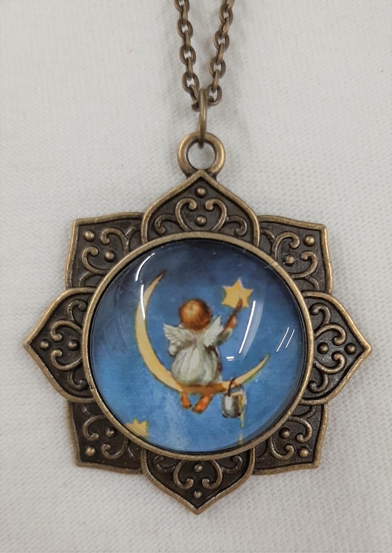 Peut inclure: Un collier de couleur bronze avec un pendentif sur le th&egrave;me c&eacute;leste. Le pendentif pr&eacute;sente une image ronde d'un ange sur un croissant de lune tendant la main vers une &eacute;toile sur fond de ciel bleu. Le pendentif est orn&eacute; d'un cadre floral.