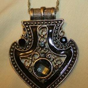 Puede incluir: Un collar con un colgante de plata, con un diseño ornamentado y esmaltado en negro. El colgante presenta una gran piedra azul facetada en el centro.