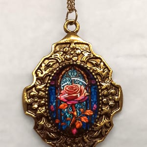 May include: A gold-toned pendant necklace with a stained-glass effect depicting a red rose. The pendant has an ornate, floral-patterned frame and hangs from a fine chain. The rose has red petals, green leaves, and is set against a blue background.