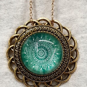 May include: A gold-colored pendant necklace featuring a round, green glass cabochon with a white mandala design. The pendant is surrounded by a decorative gold-tone frame and hangs from a delicate gold chain. The necklace is a bohemian style.