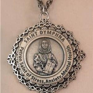 Puede incluir: Collar con colgante plateado que representa a Santa Dymphna, patrona de la salud mental, el estrés y la ansiedad. El colgante tiene un marco calado adornado e incluye el texto "Saint Dymphna" y "Patrona del estrés, la ansiedad, la salud mental."