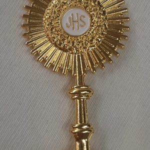 Puede incluir: Una custodia dorada con un diseño de rayos de sol. El elemento central presenta un círculo blanco con las letras "JHS". La custodia tiene una cruz en la parte superior y una base decorativa.