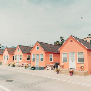 May include: A row of five pastel pink beach houses with white trim and red tile roofs. The houses have white doors and windows. There are potted plants and outdoor furniture in front of the houses.