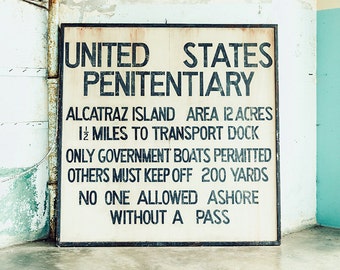 May include: A black and white sign with the text "UNITED STATES PENITENTIARY" and information about Alcatraz Island, including the area, distance to the transport dock, and rules for visitors. The sign is mounted on a white wall with a rusty pipe running vertically along the left side.