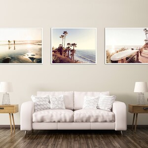 May include: Three framed photographs of a beach scene. The first photo shows two people walking on the beach at sunset. The second photo shows palm trees and a beach with waves crashing on the shore. The third photo shows a surfboard propped up on a wooden railing.