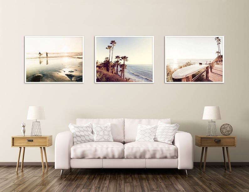 May include: Three framed photographs of a beach scene. The first photo shows two people walking on the beach at sunset. The second photo shows palm trees and a beach with waves crashing on the shore. The third photo shows a surfboard propped up on a wooden railing.