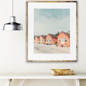 May include: A framed photograph of a row of five pastel pink beach houses with white trim and doors. The houses are lined up on a street with a light blue sky and a few clouds in the background.