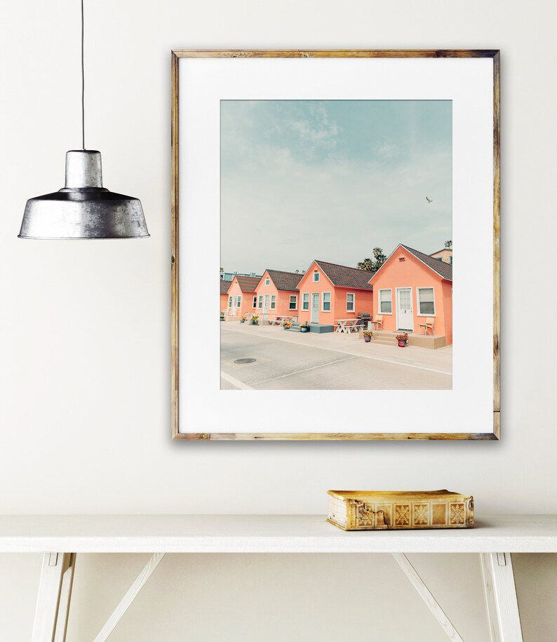 May include: A framed photograph of a row of five pastel pink beach houses with white trim and doors. The houses are lined up on a street with a light blue sky and a few clouds in the background.