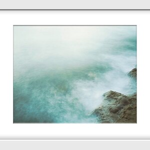 May include: A misty, ethereal seascape with soft, swirling waves crashing against a rocky shore. The water is a pale turquoise color, and the sky is a hazy white.