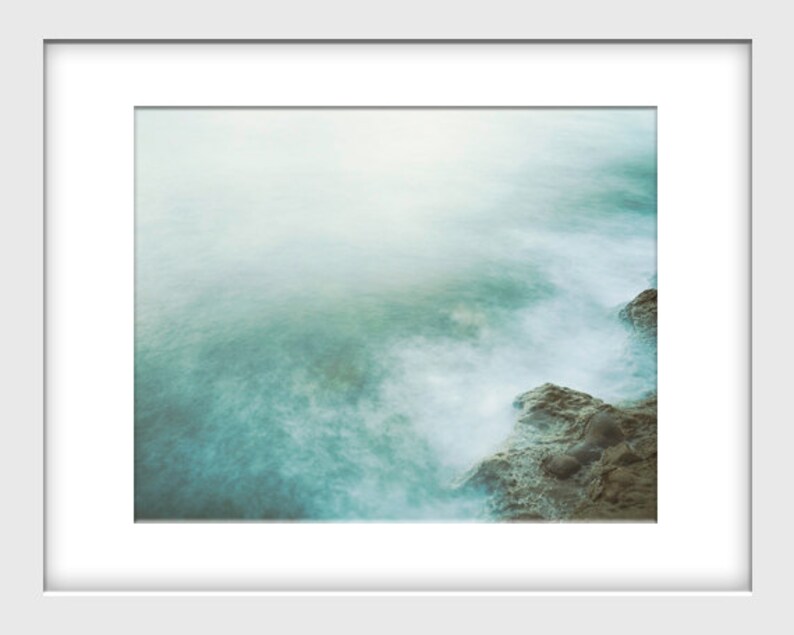 May include: A misty, ethereal seascape with soft, swirling waves crashing against a rocky shore. The water is a pale turquoise color, and the sky is a hazy white.