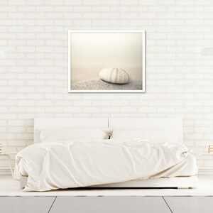 May include: A white bed with white bedding and a white frame. A framed photograph of a white sea urchin on a sandy beach hangs above the bed. The wall behind the bed is white brick.