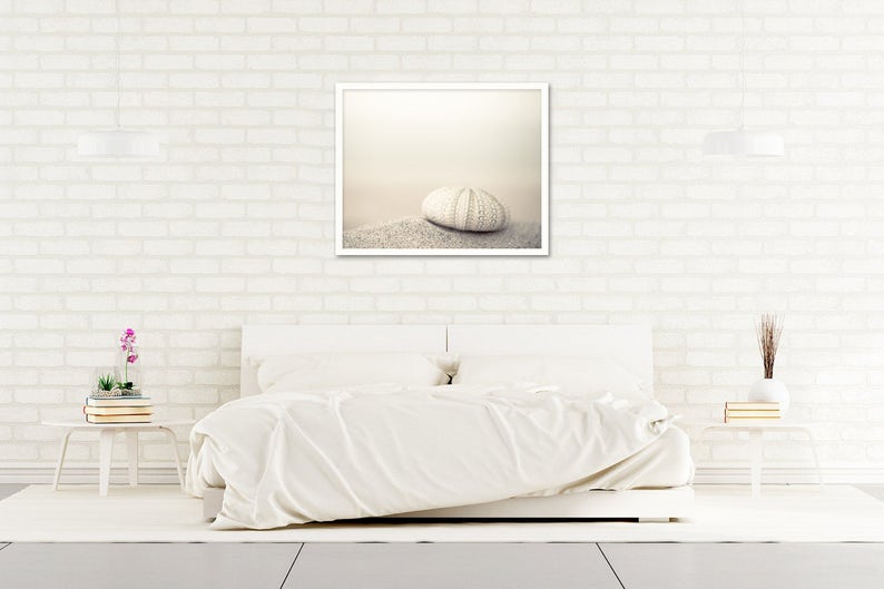 May include: A white bed with white bedding and a white frame. A framed photograph of a white sea urchin on a sandy beach hangs above the bed. The wall behind the bed is white brick.