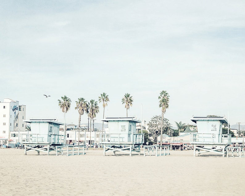 May include: A beach scene with three lifeguard stands on a sandy beach. The stands are painted light blue and have a wooden frame. There are palm trees in the background and a clear blue sky.