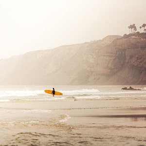 May include: A lone surfer walks out of the water with a yellow surfboard in hand, against a backdrop of a misty cliffside with a few palm trees at the top.