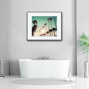 Pu&ograve; includere: Una fotografia incorniciata di palme in silhouette contro un cielo verde-azzurro con un sole brillante che splende attraverso le foglie. La fotografia &egrave; appesa a un muro bianco sopra una vasca da bagno bianca.