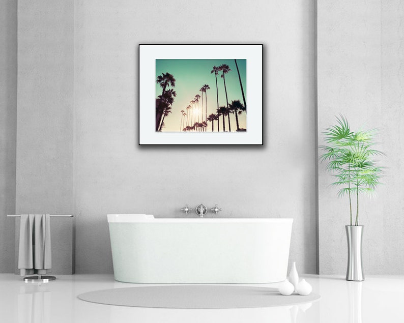 Pu&ograve; includere: Una fotografia incorniciata di palme in silhouette contro un cielo verde-azzurro con un sole brillante che splende attraverso le foglie. La fotografia &egrave; appesa a un muro bianco sopra una vasca da bagno bianca.