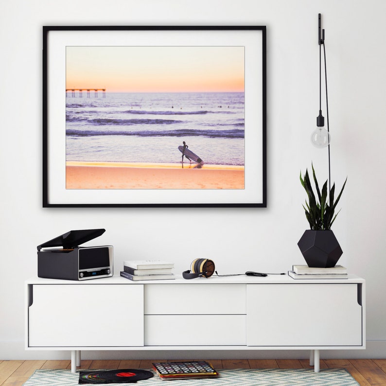May include: A framed photograph of a surfer walking on a beach with a surfboard under their arm. The beach is sandy and the water is blue. The sky is pink and orange. The photo is hanging on a white wall above a white entertainment center with a record player, headphones, and books.