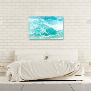 May include: A turquoise blue ocean wave crashing against the shore, creating a foamy white crest. The wave is captured in a photograph and displayed on a white brick wall above a white bed.