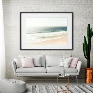May include: A white couch with two pink pillows sits in front of a framed photograph of a beach scene. A small side table with a glass vase and a cactus plant in a terracotta pot are also in the room.