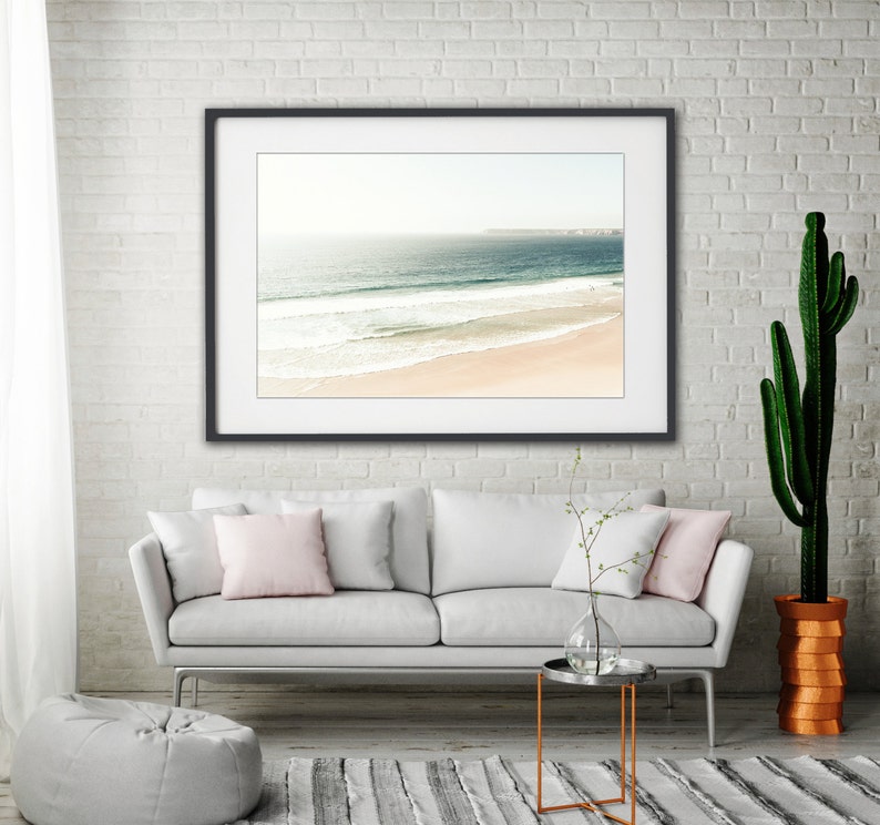 May include: A white couch with two pink pillows sits in front of a framed photograph of a beach scene. A small side table with a glass vase and a cactus plant in a terracotta pot are also in the room.