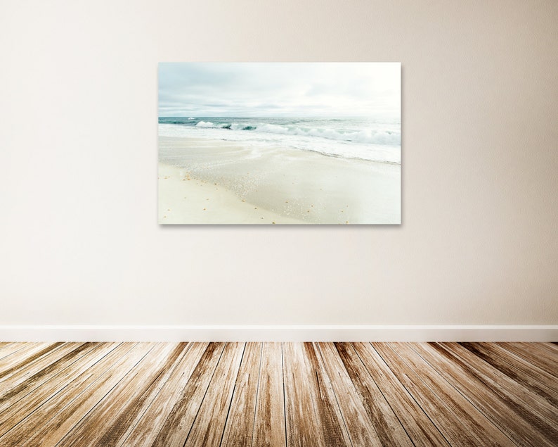 May include: A photograph of a beach scene with white sand and blue waves crashing on the shore. The sky is a light blue with some clouds.