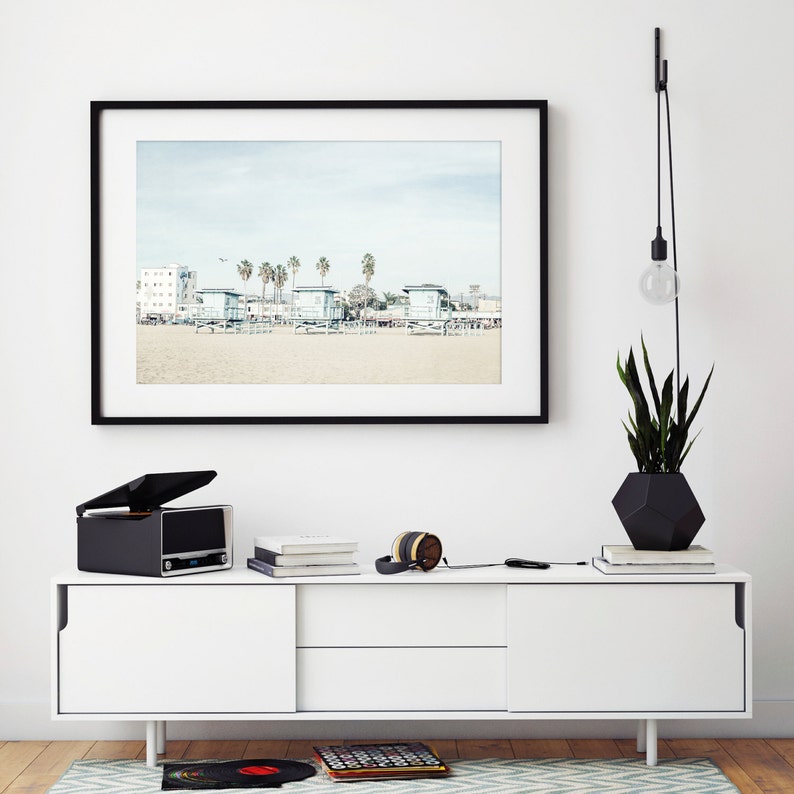 May include: A white media console with two drawers and a black record player on top. The console is in front of a white wall with a framed photograph of a beach scene and a black pendant light hanging from the ceiling.