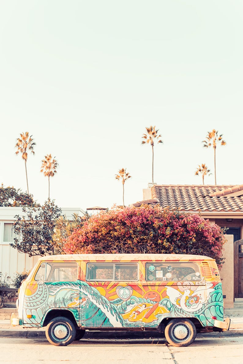 May include: A vintage yellow van with colorful psychedelic artwork featuring a peace sign, a sun, and waves. The van is parked on a street with palm trees in the background.