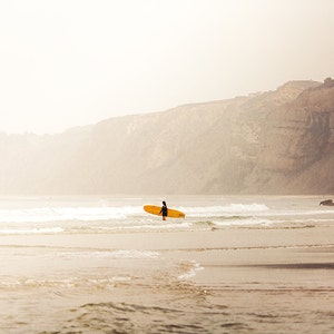 May include: A lone surfer stands in the shallows of a beach, holding a yellow surfboard. The beach is sandy and the water is a light blue. In the background, a cliff rises up from the beach, with a few trees growing on top.