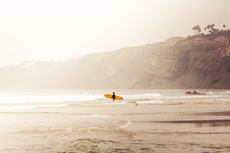 May include: A lone surfer stands in the shallows of a beach, holding a yellow surfboard. The beach is sandy and the water is a light blue. In the background, a cliff rises up from the beach, with a few trees growing on top.