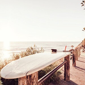 May include: A white surfboard resting on a wooden railing overlooking a beach with a blue ocean and a clear sky. The surfboard is wet and has a leash attached to it.