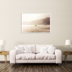 May include: A framed photograph of a surfer paddling out on a surfboard in the ocean. The photo is taken from the beach, looking out towards the water. The surfer is silhouetted against the setting sun, and the water is a deep blue.