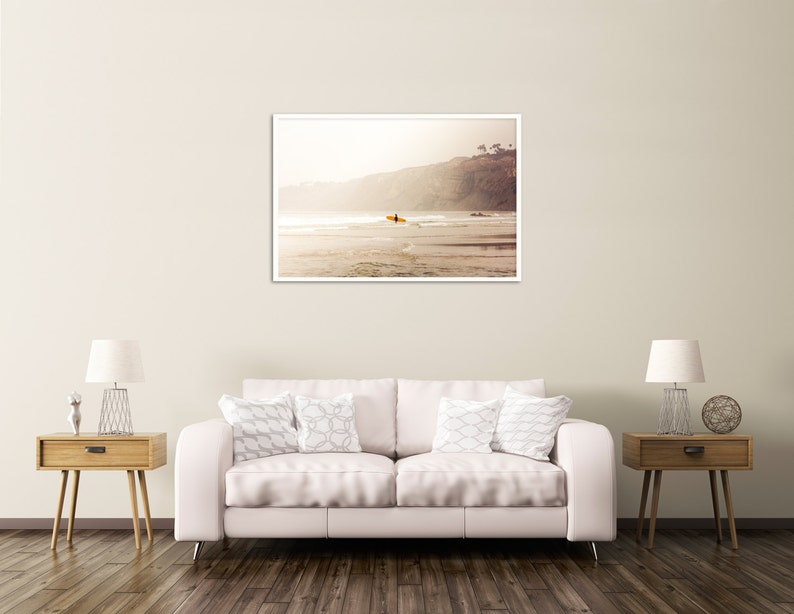May include: A framed photograph of a surfer paddling out on a surfboard in the ocean. The photo is taken from the beach, looking out towards the water. The surfer is silhouetted against the setting sun, and the water is a deep blue.
