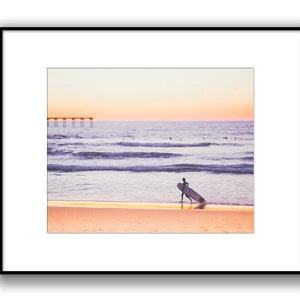 May include: A lone surfer walks along a sandy beach with a surfboard under their arm, facing away from the camera. The ocean is in the background with a pier in the distance. The sky is a soft pink and orange color, suggesting a sunset.