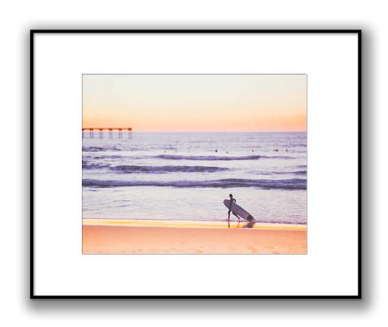 May include: A lone surfer walks along a sandy beach with a surfboard under their arm, facing away from the camera. The ocean is in the background with a pier in the distance. The sky is a soft pink and orange color, suggesting a sunset.