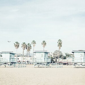 May include: A beach scene with four lifeguard stands on a sandy beach. The stands are painted light blue and have a white roof. There are palm trees in the background and a building in the distance.