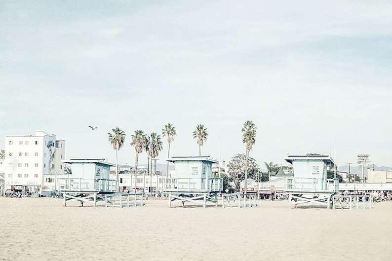 May include: A beach scene with four lifeguard stands on a sandy beach. The stands are painted light blue and have a white roof. There are palm trees in the background and a building in the distance.