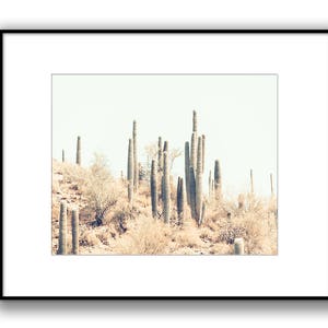 Pu&ograve; includere: Stampa incorniciata con un paesaggio desertico in tonalit&agrave; seppia. L'immagine mostra un gruppo di alti cactus saguaro e vegetazione desertica contro un cielo chiaro. L'opera d'arte &egrave; presentata in una cornice nera con un passe-partout bianco.