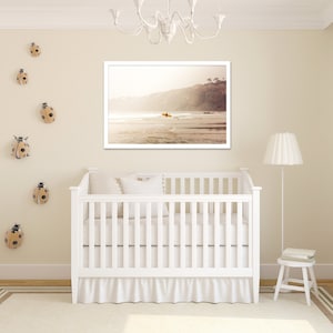 May include: A white crib with a white ruffle skirt, a white pillow, and a framed photograph of a beach scene with a surfer riding a wave. The wall is decorated with ladybug figurines.