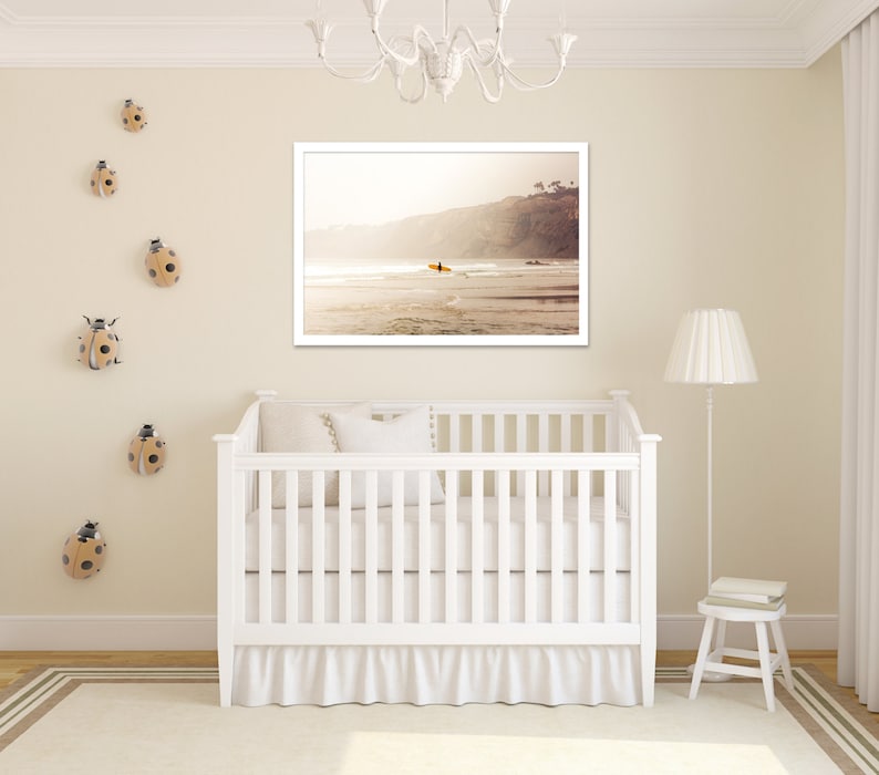 May include: A white crib with a white ruffle skirt, a white pillow, and a framed photograph of a beach scene with a surfer riding a wave. The wall is decorated with ladybug figurines.