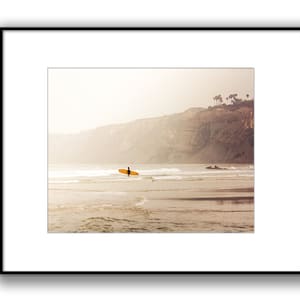 May include: A lone surfer walks along a sandy beach with a surfboard in hand, with a cliff and a few trees in the background. The sky is a hazy, pale yellow.