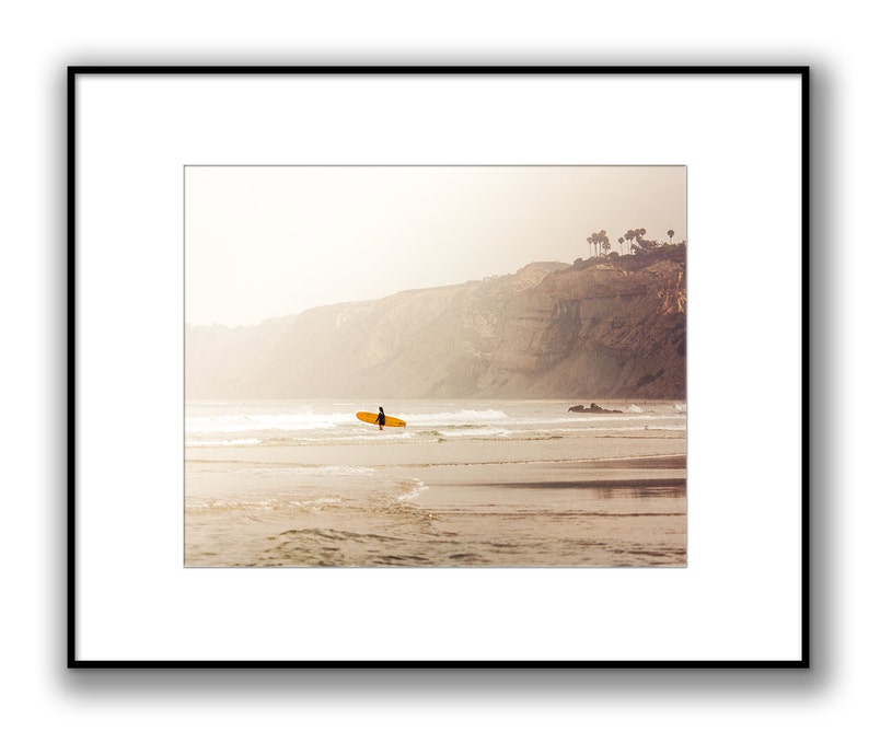 May include: A lone surfer walks along a sandy beach with a surfboard in hand, with a cliff and a few trees in the background. The sky is a hazy, pale yellow.