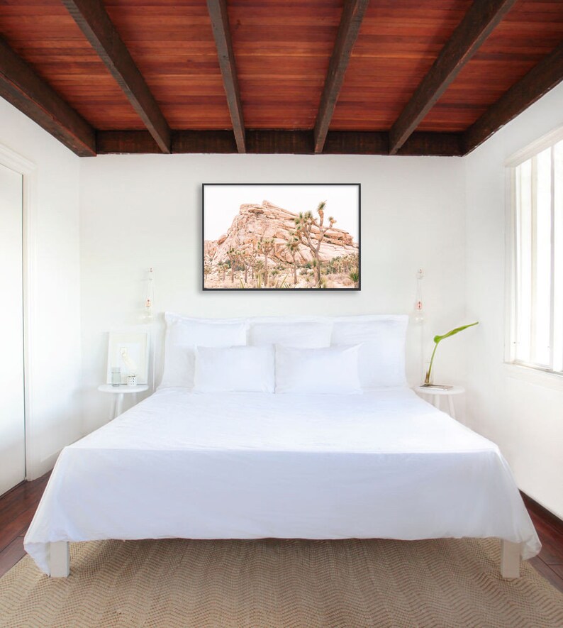 May include: A white bed with white bedding and pillows in a bedroom with a wood ceiling. A framed photograph of a desert landscape with a rocky mountain and Joshua trees hangs above the bed.