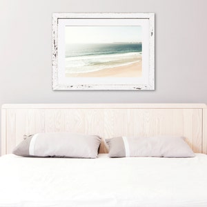May include: A white wooden bed frame with a white headboard and two white pillows. A framed photograph of a beach scene hangs on the wall above the headboard.