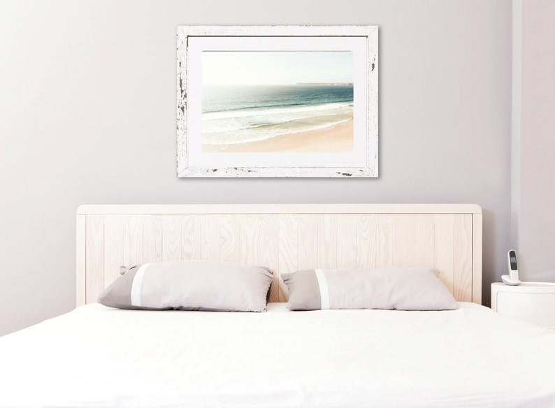 May include: A white wooden bed frame with a white headboard and two white pillows. A framed photograph of a beach scene hangs on the wall above the headboard.
