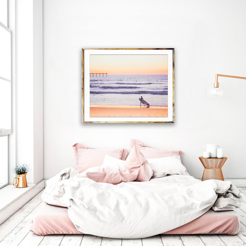 May include: A framed photograph of a surfer walking on a sandy beach with a surfboard under their arm. The ocean is in the background with a pier in the distance. The sky is a vibrant orange and pink.