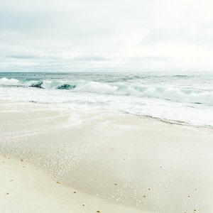 May include: A white sandy beach with foamy waves crashing on the shore. The sky is overcast and the water is a light blue.
