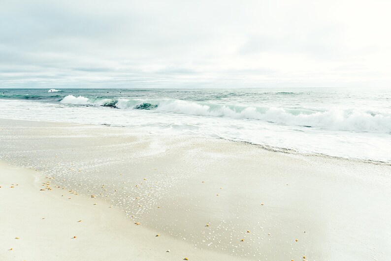 May include: A white sandy beach with foamy waves crashing on the shore. The sky is overcast and the water is a light blue.