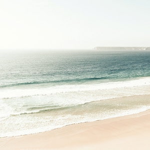 May include: A panoramic view of a sandy beach with white foamy waves crashing on the shore. The ocean is a light blue color and the sky is a hazy white.