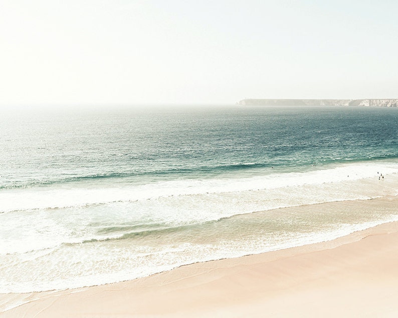 May include: A panoramic view of a sandy beach with white foamy waves crashing on the shore. The ocean is a light blue color and the sky is a hazy white.